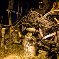 A cluttered, rustic workshop with various tools and metal parts. In the foreground, a stationary bicycle wheel is mounted on a log. Nearby, an old grinder and other metalworking tools are placed on tree stumps. The background shows an old guitar and various hanging items from the Give and Get festival 2013 in a dimly lit space.
