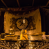 A dimly lit stage adorned with various musical instruments and equipment, reminiscent of the 2013 festival. A guitar hangs in the center, with a mandolin resting on the table below it. Two speakers and several electronic devices are arranged on either side. A flag with a lion crest hangs behind, and tribal patterns decorate the table.