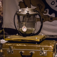 An antique miner's lamp with a glass encasement and metal top is displayed atop a worn, brown leather suitcase. The lamp has a small emblem attached to it. The background features a partially visible banner with a stylized lion design from the 2013 Give and Get festival. The scene is dimly lit.