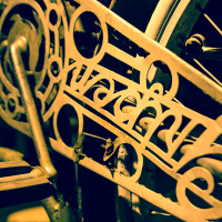 Close-up view of a vintage bicycle crank and chain guard with intricate geometric cutouts. The metal parts have a weathered, golden patina, reminiscent of the decor at the 2013 Give and Get event. The image features an artistic, slightly faded look, highlighting the detailed craftsmanship of the old bicycle components.