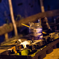 A dimly lit image of a workspace featuring a plastic paint palette with several compartments filled with various shades of paint, remnants from the 2013 Give and Get festival. A partially used paintbrush rests atop the palette. In the background, an open jar labeled "SUDRABA" contains liquid, with a yellow paintbrush jutting out.