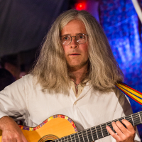 At the 2013 Give and Get festival, a person with long gray hair and glasses is playing an acoustic guitar decorated with colorful patterns around the sound hole. They are wearing a white shirt and have a serious expression. The background is dimly lit with blue and red lighting, creating a vibrant atmosphere.