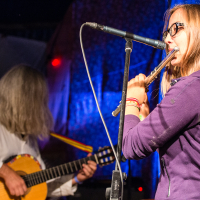 A young woman in glasses and a purple hoodie plays the flute into a microphone on stage at the 2013 Give and Get festival. In the background, an older person with long gray hair plays a guitar. The scene is lit with blue and purple stage lights, suggesting a live musical performance.