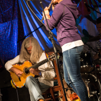 At the Give and Get festival 2013, a woman wearing a purple hoodie and jeans plays a flute into a microphone on stage. Beside her, a man with long white hair in a white shirt and jeans strums a yellow acoustic guitar. The background is decorated with colorful fabric, and a drum set is visible behind them.