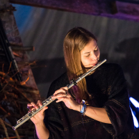 A person with long, light brown hair plays a flute, wearing a black sweater and standing in a dimly lit environment. The background features wooden beams and soft lighting, creating a cozy atmosphere reminiscent of the Give and Get festival. The person appears focused and passionate about their music.