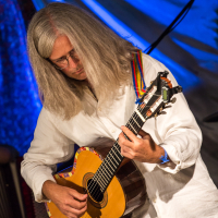 At the 2013 festival, a person with long gray hair and glasses plays an acoustic guitar. Dressed in a white shirt and using a multicolored strap, they perform against the blue-lit drapery backdrop, adding colorful ambiance. The musician appears deeply focused and absorbed in the music.
