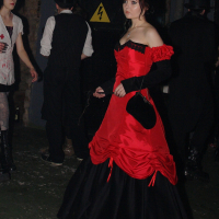 A woman in a vivid red and black Victorian-style gown stands in a dimly lit room, reminiscent of Elektra '09, holding a black fan. She has fair skin and dark hair styled in an updo. Other people in the background wear dark, gothic attire, including top hats and suits, setting the scene for an eerie Halloween party amidst exposed brick and caution signs.
