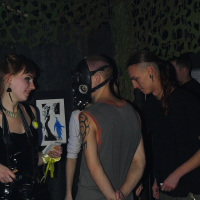 A group of four people are seen interacting in a dimly lit room with dark walls and camouflage netting in the background, reminiscent of an Elektra '09 Halloween party. One person wears a sleeveless shirt and a gas mask, while another has long hair pulled back and wears a black shirt. A woman dressed in a shiny black outfit holds a framed picture.
