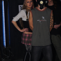 Three people, one wearing a gas mask, stand in a dimly-lit room at a Halloween party. The person in front wears a sleeveless shirt, revealing a tattooed arm reminiscent of Elektra '09. Behind, a woman in a pink plaid skirt and black top leans on an industrial-style lamp emitting blue light. Another person, partially obscured, stands in the background.