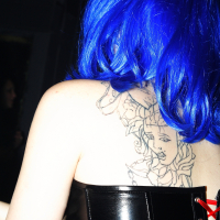 A person with bright blue hair wearing a black patent leather corset with red lacing is shown from the back at what appears to be a Halloween party. Their upper back displays a detailed tattoo of a woman's face surrounded by intricate designs, reminiscent of Elektra '09. The image is taken in a dimly lit setting.
