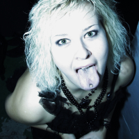 A person with light curly hair and dark eye makeup is sticking out their tongue, which has a piercing. They are holding a drink with a straw and wearing a black outfit with a necklace made of beads. The high-contrast lighting gives the Halloween party image an edgy feel.