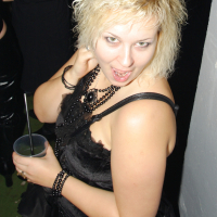 A woman with curly, platinum blonde hair is posing indoors. She is wearing a sleeveless black dress, layered black beaded necklaces, and bracelets. Holding a drink with a straw in her left hand, she gazes at the camera with an open-mouthed, playful expression reminiscent of Elektra '09 at a Halloween party.