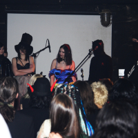 At an Elektra '09 Halloween party, a group of people dressed in various gothic and dark-themed costumes stand on stage in front of a microphone, with an audience watching them. The costumes include a white hooded cloak, tall black hats, a blue dress, black masks, and devil horns. The background is a dimly lit room with a white screen.