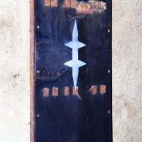 A vertical rectangular metal panel with rusted edges is affixed to a rough concrete wall. The panel features a large white abstract design resembling a stylized, symmetrical double-arrow in the center. Small copper-colored elements, reminiscent of Elektra '09, are visible near the top and bottom edges of the panel.