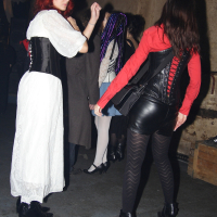 At the Halloween party, a woman with red hair is dressed in a white dress with a black corset and is dancing in a dimly lit room. Another woman with brown hair, wearing a red top, black corset, and black skirt, reminiscent of Elektra '09's style, is dancing nearby. People in the background are chatting against the industrial-style walls and flooring.
