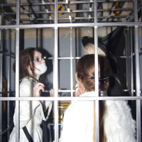 Three people are standing inside a metal cage at the Halloween party. The person on the left wears glasses, a white outfit, a mask, and carries a black bag. The person in the center, with long hair, is seen from the back wearing a white feathered garment reminiscent of Elektra '09. The person on the right wears a tall witch's hat.
