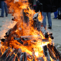 A massive bonfire ignites amid a bustling crowd.