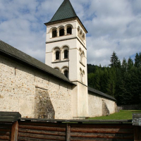 An imposing white bell tower with a conical roof stands adjacent to a solid stone church in Romania, flanked by a verdant forest under a blue sky with scattered clouds, enclosed by a rustic wooden