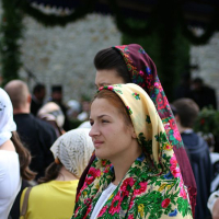 A woman in a vibrant floral headscarf attends a cultural or religious gathering in Romania, her expression contemplative, amidst a crowd of similarly dressed individuals.