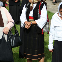A woman, clad in traditional Romanian attire with a patterned headscarf, embroidered vest, and striped skirt, stands amidst a gathering, her hands clasped together, showcasing cultural heritage and attire.