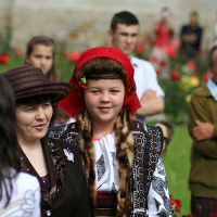 A young individual in traditional Romanian embroidered attire, including a red headscarf, confidently walks alongside a woman wearing a brown hat, as others gather amidst a vibrant floral backdrop, hinting at a cultural