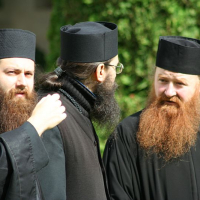 Three orthodox christian clergy members from Romania in traditional black vestments and caps engage in a conversation, with two of them facing the camera showcasing their distinct beards while the third, with his back turned,