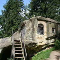 A rustic wooden staircase leads up to an old, stone hermitage built into a large boulder in Romania, surrounded by lush greenery and tall trees under a clear blue sky.
