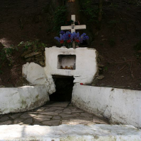 A secluded forest shrine in Romania with a white fountain structure, adorned with crosses and vibrant blue flowers, offering a tranquil spot for reflection amidst the trees.