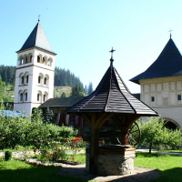 A serene monastery garden in Romania with a wooden gazebo, surrounded by lush trees and flowers, flanked by the monastery's bell tower and gatehouse on a bright sunny day.