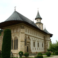 An old, ornate stone church with a conical roof and a tall bell tower, surrounded by manicured hedges and blooming flowers, stands against a cloudy sky in Romania.