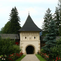 A tranquil pathway leads to a traditional stone gatehouse with a pointed roof, surrounded by lush greenery and vibrant red roses, evoking a serene, historical ambiance in Romania.