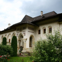 An elegant traditional monastery in Romania with an ornate porch, surrounded by a manicured lawn and blooming roses under a cloudy sky.