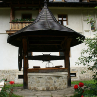 A traditional wooden well with a shingled roof, featuring a wrought iron crank, stands in the courtyard of an elegant Romania-style house with decorative eaves, framed by blooming red roses.