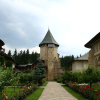 A serene pathway lined with blooming flowers leads to an ancient stone tower in Romania under a cloudy sky, suggestive of a rich history nestled within a peaceful garden.