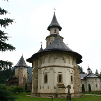 A historic church with a characteristic spire, clad in white, stands amidst greenery, with an old fortification tower visible in the background, illustrating an ancient Romanian architectural ensemble in a tranquil, forest