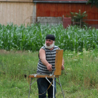 An artist in a striped tank top and black headband stands by an easel, painting en plein air amidst rural Romanian scenery with greenery and a red-roofed building in the backdrop.