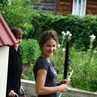 A smiling young woman in casual attire turns towards the camera while an older person leans against a wall in the background, with a quaint wooden cabin and lush greenery setting a serene backdrop in Romania.