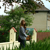A woman stands by a white fence with birdhouses, holding a painting, amidst lush greenery with a residential home in the background in Romania. She seems to be either placing the artwork or admiring