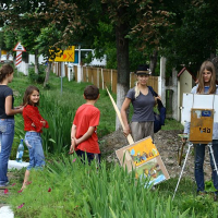 A group of children stand and watch a young artist painting outdoors by a roadside in Romania. The artist, wearing glasses and a hat, works on a landscape on an easel. They stand near a grassy area with a wooden fence and street signs in the background. The scene is relaxed, inviting travelers to enjoy local art tourism.