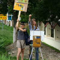 Two smiling artists in Romania outdoors with easels, one standing painting on a canvas, while the other showcases finished artwork.