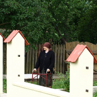 A person stands between two miniature houses, holding onto a red and white barrier, surrounded by lush greenery in what appears to be a park or outdoor exhibit in Romania, evoking a whimsical,