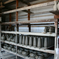 Rows of unfinished pottery pieces, including vases and bowls, sit on wooden shelves to dry in a Romanian workshop environment, showcasing various stages of the ceramic process.