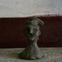 A handcrafted clay bust with distinct facial features sits on a wooden surface against a wall with peeling paint in Romania, accompanied by a perched butterfly on its head.