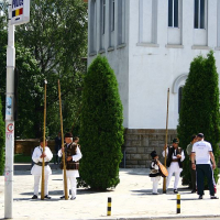 Individuals in traditional attire stand near a street sign in Bucharest, engaging in conversation, while others walk by on a sunny day with greenery and a white building in the background.