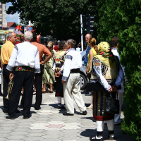 People in traditional Romanian attire gather on a sunny day, possibly for a cultural event or festival, marked by a convivial atmosphere amidst the urban surroundings.