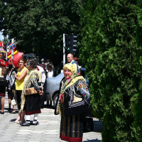 A woman dressed in traditional Romanian folk attire, including a patterned shawl and skirt, stands on a sunny street, with trees and a festive atmosphere in the background.