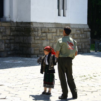 A child dressed in traditional Romanian folk attire walks alongside an adult in a military-style uniform, possibly during a cultural event or ceremony, in a courtyard against the backdrop of a white building with arched windows