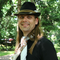 A smiling man with long hair wearing a black t-shirt, a patterned scarf, and a fedora hat poses casually in a lush, green park setting in Romania.