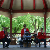 A quartet of musicians dressed in red and black perform under a gazebo in Romania, with trees in the background, featuring a trombone, drums, accordion, and trumpet.