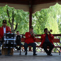 A brass band, dressed in traditional uniforms with red jackets and black hats, enthusiastically performs on a wooden gazebo amidst lush greenery in Romania, with a percussionist keeping the rhythm on a snare